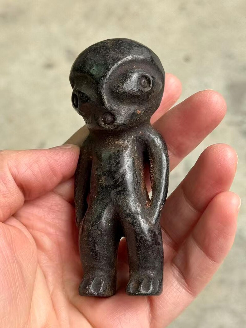 May include: A small, dark-colored figurine of a humanoid figure. The figure has a rounded head, large eyes, and a simple body with arms at its sides. The figurine is held in a hand.