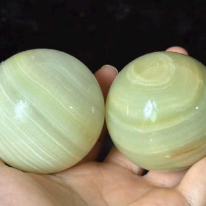 May include: Two polished, spherical stones with a smooth, glossy finish. The stones are light green in colour and feature distinct, swirling bands of translucent and opaque white and pale yellow mineral layers throughout their rounded surfaces.