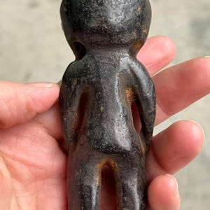 May include: A small, dark-colored figurine in the shape of a stylized human figure. The sculpture has a rounded head, a simple body with open spaces for arms and legs, and a textured surface. The figurine is held in a hand.