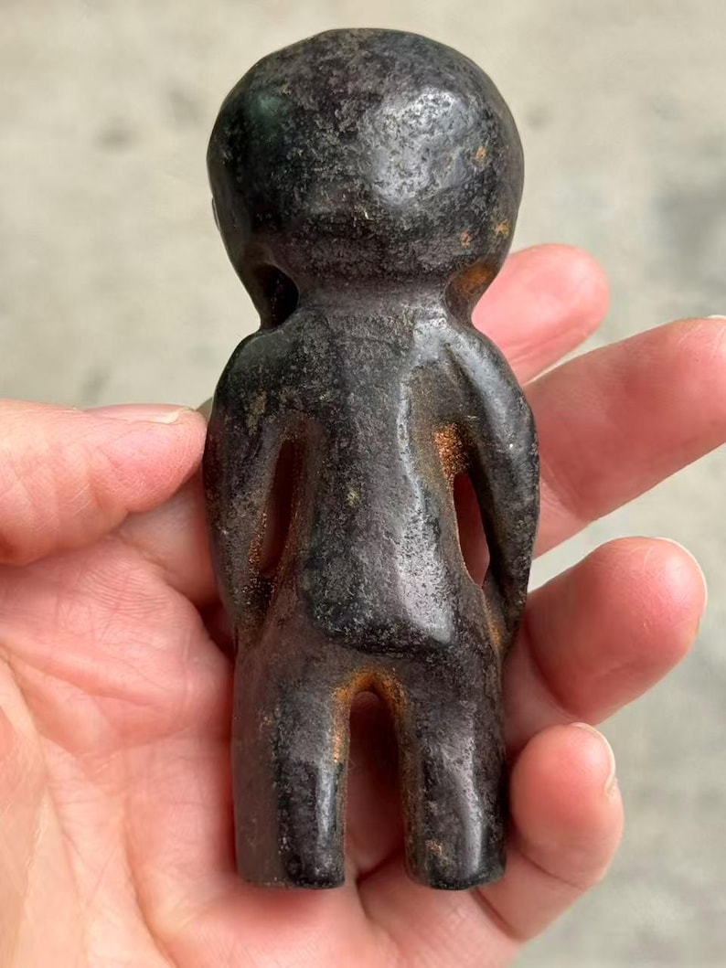 May include: A small, dark-colored figurine in the shape of a stylized human figure. The sculpture has a rounded head, a simple body with open spaces for arms and legs, and a textured surface. The figurine is held in a hand.