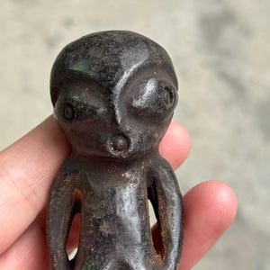 May include: A dark brown, stylized figurine with a rounded head, large eyes, and a small mouth. The figure has a simple, humanoid form with open arm holes. The surface has a textured, aged appearance.