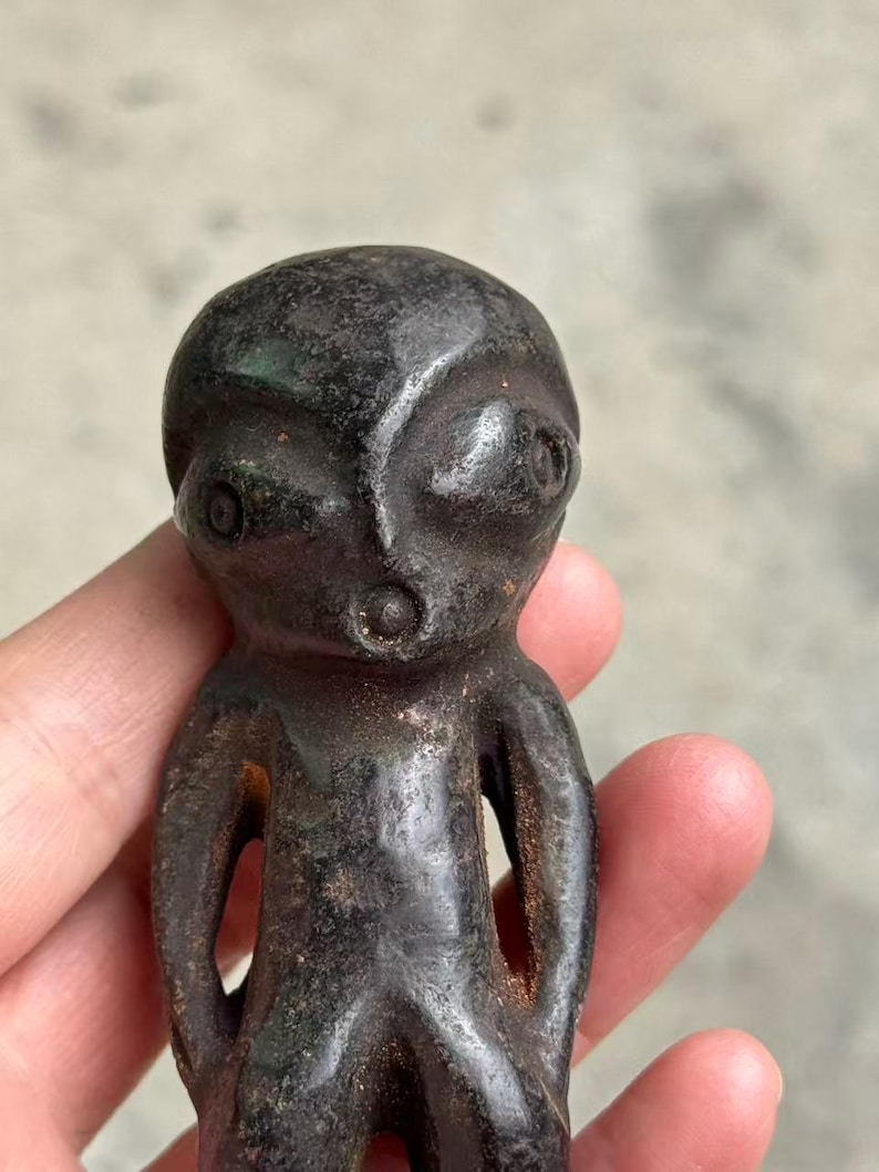 May include: A dark brown, stylized figurine with a rounded head, large eyes, and a small mouth. The figure has a simple, humanoid form with open arm holes. The surface has a textured, aged appearance.