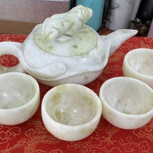 Puede incluir: Juego de té de jade blanco con una tetera y cuatro tazas a juego. La tetera tiene una tapa y un pico en forma de rana. Las tazas y la tetera están sobre una superficie roja estampada. Un artículo decorativo.
