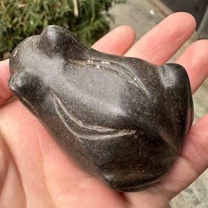 May include: A dark gray, carved stone frog figurine. The frog is in a resting position with etched details. The sculpture is held in a hand, showcasing its size and texture. A decorative item.