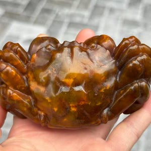 May include: A detailed, amber-colored crab sculpture. The crab is carved with intricate details, showcasing its claws, body, and legs. The sculpture has a polished, glossy finish, and is held in a hand.