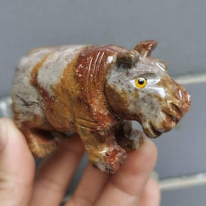 Puede incluir: Figura de piedra tallada a mano de un hipopótamo. La escultura presenta una mezcla de tonos marrones, beige y grises, con ojos amarillos. El hipopótamo se representa en posición agachada, mostrando los detalles del tallado en piedra.