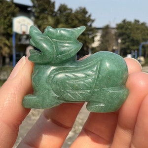 May include: A carved green jade figurine of a mythical creature, possibly a Pixiu or similar guardian beast. The figurine is detailed with etched patterns and is held in a hand against a blurred outdoor background. The jade has a polished finish.