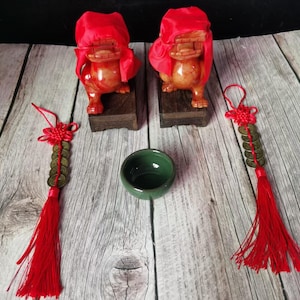 Puede incluir: Dos estatuas de rana de color naranja-marrón con cubiertas de tela roja, sobre bases de madera, flanquean un pequeño cuenco verde. Dos adornos de borlas rojas con monedas están a cada lado. Los artículos están dispuestos sobre una superficie de vetas de madera clara.