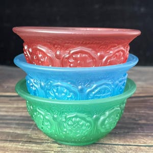 May include: A stack of three translucent glass bowls in red, blue, and green. Each bowl features a repeating embossed floral pattern around the exterior, a wide flared rim, and a smooth interior surface, creating a vibrant, layered appearance.