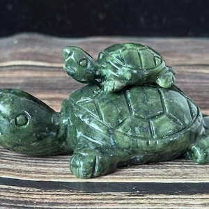 May include: A carved green stone turtle figurine with a smaller turtle on its back. The turtles have detailed shell patterns and are set against a wooden background. The figurine is approximately 10 cm long.