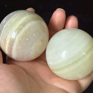 Può includere: Due pietre sferiche levigate con superfici lisce. Una sfera presenta bande traslucide bianche e verde pallido con sottili inclusioni color marrone chiaro, mentre l'altra sfera mostra un colore verde menta chiaro uniforme con lievi striature bianche orizzontali.