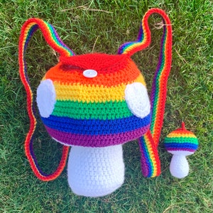 May include: A rainbow crocheted mushroom backpack with a smaller matching mushroom charm. The backpack has a white crocheted base and a red, orange, yellow, green, blue, indigo, and violet crocheted top.