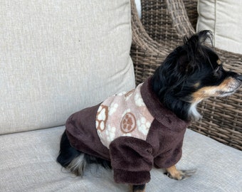 May include: A small brown and tan dog wearing a brown fleece sweater with paw print designs. The dog is sitting on a light brown couch.