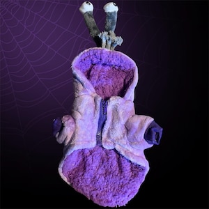May include: Purple plush dog costume with a zip closure and a hood. The costume has two large, grey eyes on top of the hood.