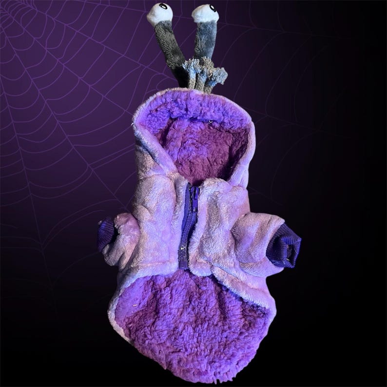 May include: Purple plush dog costume with a zip closure and a hood. The costume has two large, grey eyes on top of the hood.