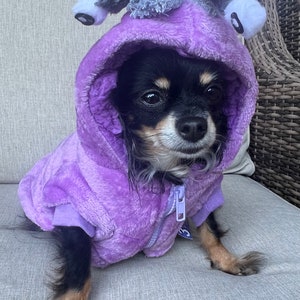 May include: A small black and brown chihuahua wearing a purple hooded sweater with a grey and white plush toy on top of the hood. The dog is sitting on a grey sofa.
