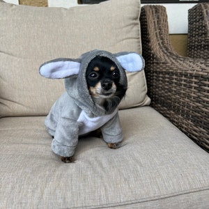 May include: A small black and brown dog wearing a gray hooded animal costume with large ears. The dog is sitting on a light gray couch.