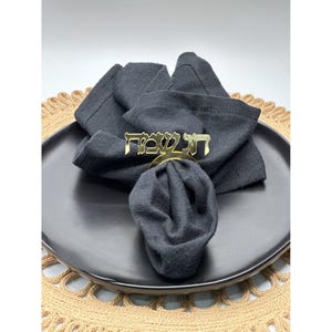 May include: A black linen napkin is held by a gold-colored napkin ring with Hebrew text. The napkin rests on a black plate, which is set on a woven placemat.