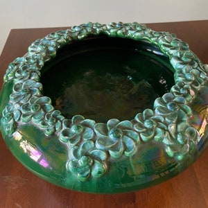 Puede incluir: Un cuenco grande, redondo, de cerámica verde con un diseño floral en relieve alrededor del borde. El cuenco tiene un acabado brillante y un ligero brillo iridiscente.