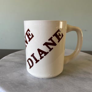 May include: Cream-colored ceramic mug with the name "DIANE" printed in diagonal, dark red letters. The mug has a curved handle and is set on a marble surface.