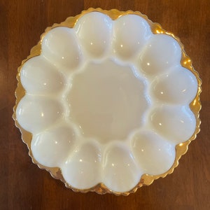 Op de afbeelding: Een ronde serveerschaal van wit melkglas voor gevulde eieren met twaalf ovale inkepingen in een cirkel rond een glad midden, afgewerkt met een geschulpte rand voorzien van een metallic gouden bies.