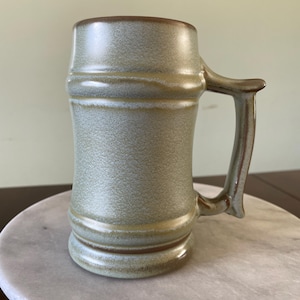 May include: A stoneware mug with a textured, light green body and a brown rim and handle. The mug has a cylindrical shape with a sturdy handle, perfect for enjoying hot beverages. The mug is sitting on a white marble surface.