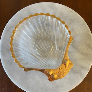 May include: A clear glass seashell dish with a gold rim and detailed gold accents. The dish is sitting on a white marble surface.