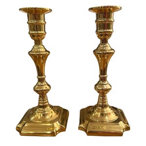 May include: Two gold-colored candlesticks with a classic design. Each candlestick has a square base, a slender stem with decorative elements, and a wide top for holding candles. The candlesticks are made of a shiny, reflective material.