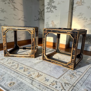 May include: Two black square side tables with gold trim and ornate floral designs. The tables are open-framed, with a hollow center. They sit on a light gray rug with a patterned border, against a wallpaper backdrop.