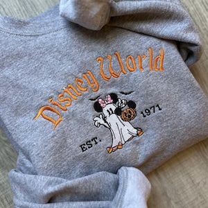 以下が含まれることがあります： オレンジ色の文字で「Disney World」と書かれたグレーのスウェットシャツ。その下に、ミニーマウスのリボンをつけたカボチャを持った漫画の幽霊がいます。「EST. 1971」の文字もスウェットシャツにあります。