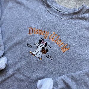以下が含まれることがあります： オレンジ色の「Disney World」の刺繍が入ったグレーのクルーネックのスウェットシャツ。カボチャのバケツを持ったミニーマウスのゴーストデザインと、その下に「EST. 1971」があります。ディズニーファン向けの、快適な長袖トップスです。