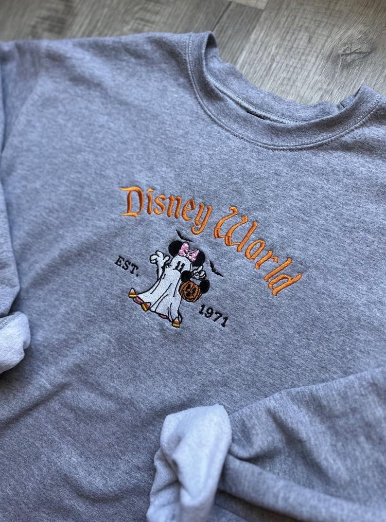 以下が含まれることがあります： オレンジ色の「Disney World」の刺繍が入ったグレーのクルーネックのスウェットシャツ。カボチャのバケツを持ったミニーマウスのゴーストデザインと、その下に「EST. 1971」があります。ディズニーファン向けの、快適な長袖トップスです。