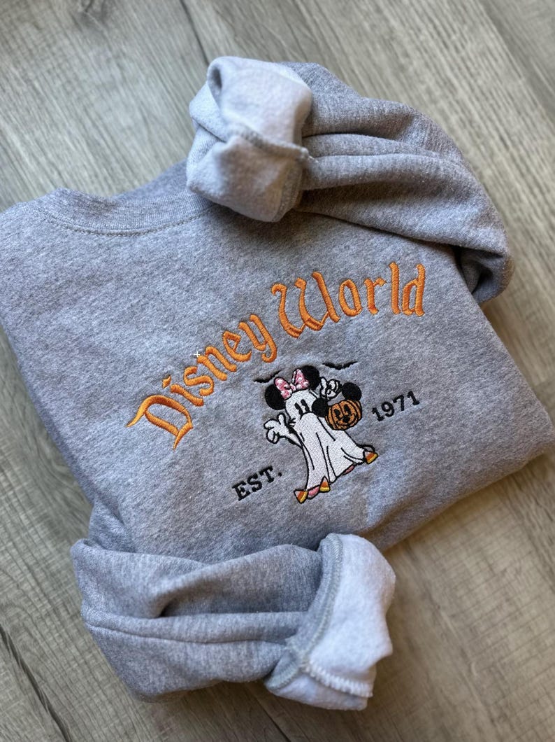 以下が含まれることがあります： オレンジ色の文字で「Disney World」と書かれたグレーのスウェットシャツ。その下に、ミニーマウスのリボンをつけたカボチャを持った漫画の幽霊がいます。「EST. 1971」の文字もスウェットシャツにあります。