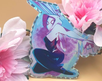 May include: A holographic sticker showing a female superhero in a dynamic pose. The character is in a black and white costume with blue and purple accents. The sticker is set against a backdrop of pink flowers.