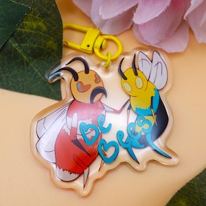 May include: A clear acrylic keychain featuring two cartoon bees. One bee is red and orange, the other is yellow and blue. The keychain has a yellow clasp and the words "be bees" in teal.