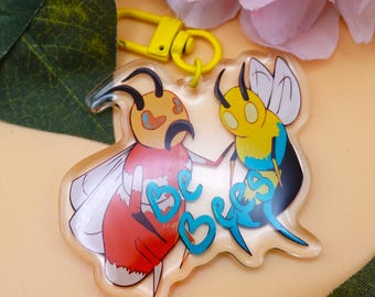 May include: A clear acrylic keychain featuring two cartoon bees. One bee is red and orange, the other is yellow and blue. The keychain has a yellow clasp and the words "be bees" in teal.