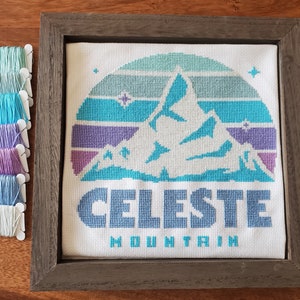May include: Cross-stitch embroidery in a wooden frame with a blue and purple mountain scene and the text "Celeste Mountain".