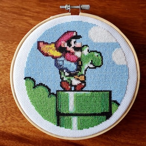 Könnte beinhalten: Eine Kreuzstich-Stickerei mit einem grünen Yoshi, der einen roten und blauen Mario auf seinem Rücken trägt. Sie stehen auf einem grünen Rohr vor einem blauen Himmel mit weißen Wolken.