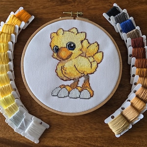 May include: A completed cross-stitch embroidery project featuring a yellow chick with black eyes. The embroidery is framed in a round wooden hoop and surrounded by spools of yellow, brown, and blue embroidery floss.