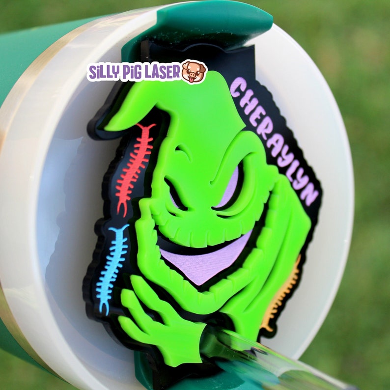 Puede incluir: Personaje Oogie Boogie impreso en 3D en verde y negro con una sonrisa morada y ciempi&eacute;s rojos, naranjas, azules y verdes. El personaje est&aacute; sobre un fondo blanco con el texto "Cherylyn" en morado y "Silly Pig Laser" en negro.