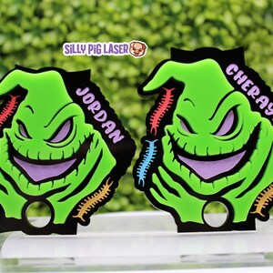 Puede incluir: Dos llaveros verdes y morados con el personaje de Oogie Boogie, con los nombres "Jordan" y "Cheraylyn" escritos en ellos. Los llaveros tienen dise&ntilde;os de ciempi&eacute;s coloridos alrededor de los bordes.