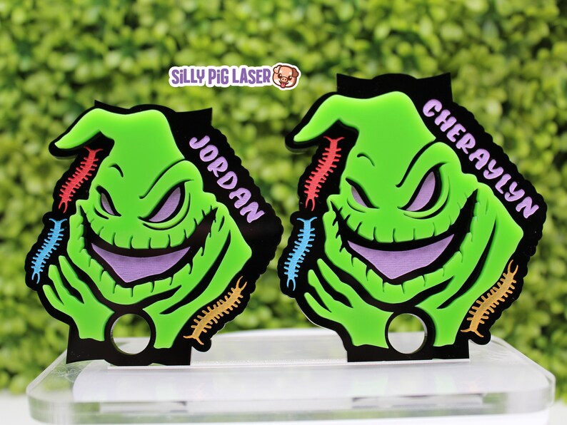 Puede incluir: Dos llaveros verdes y morados con el personaje de Oogie Boogie, con los nombres "Jordan" y "Cheraylyn" escritos en ellos. Los llaveros tienen dise&ntilde;os de ciempi&eacute;s coloridos alrededor de los bordes.