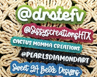 May include: A collection of glittery, colorful name tags with white text. The tags include the following names: @drstefv, @SissyscreationsHTX, CACTUS MOMMA CREATIONS, @PEARLSDIAMONDART, and Sweet GA Belle Designs. The background is a woven, beige material.