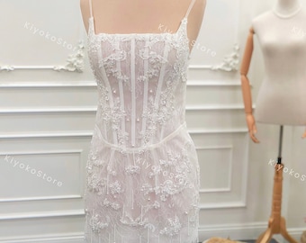 May include: A white lace mini dress with a fitted bodice and a beaded fringe hem. The dress has a square neckline and thin straps.