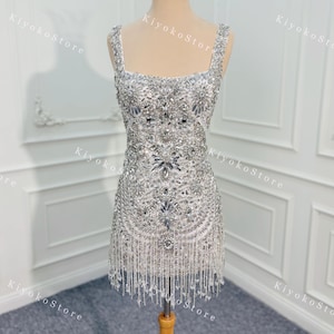 K&ouml;nnte beinhalten: Ein silbernes, perlenbesetztes Minikleid mit quadratischem Ausschnitt und Fransen. Das Kleid ist mit glitzernden Verzierungen versehen und wird auf einer Schaufensterpuppe mit goldener Krone pr&auml;sentiert. Das Kleid ist etwa 76 cm lang.
