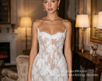 May include: A white, strapless wedding dress with intricate floral lace detailing. The short dress features a fitted bodice and a fringed hemline. The dress is designed with a sheer overlay, creating a delicate and elegant look. The KiyokoStore custom wedding dress is perfect for a bride.