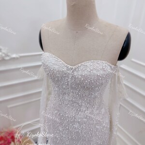 May include: A white wedding dress with a sweetheart neckline and long sleeves. The dress is covered in delicate beading and sequins.
