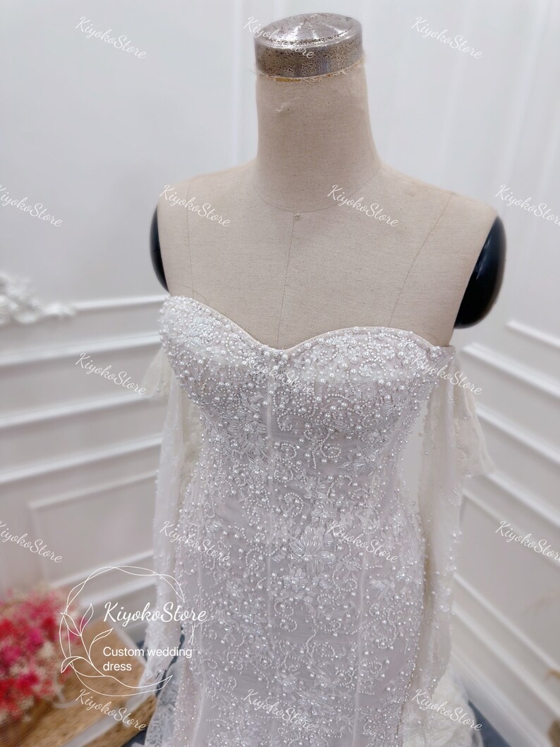May include: A white wedding dress with a sweetheart neckline and long sleeves. The dress is covered in delicate beading and sequins.