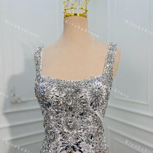 K&ouml;nnte beinhalten: Ein wei&szlig;es Abendkleid mit quadratischem Ausschnitt und mit silbernen Kristallen verziert. Eine goldene Krone sitzt auf dem Kopf der Schaufensterpuppe. Der Text "KiyokoStore" ist im Hintergrund sichtbar.