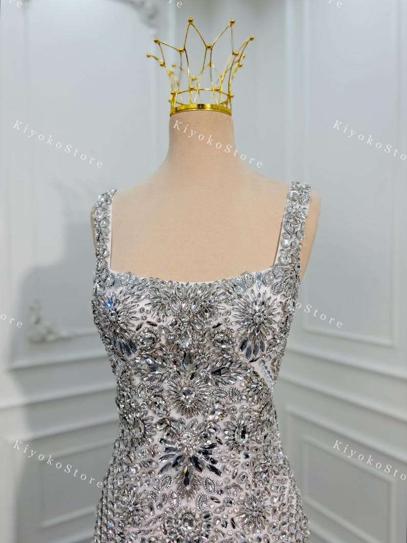 K&ouml;nnte beinhalten: Ein wei&szlig;es Abendkleid mit quadratischem Ausschnitt und mit silbernen Kristallen verziert. Eine goldene Krone sitzt auf dem Kopf der Schaufensterpuppe. Der Text "KiyokoStore" ist im Hintergrund sichtbar.